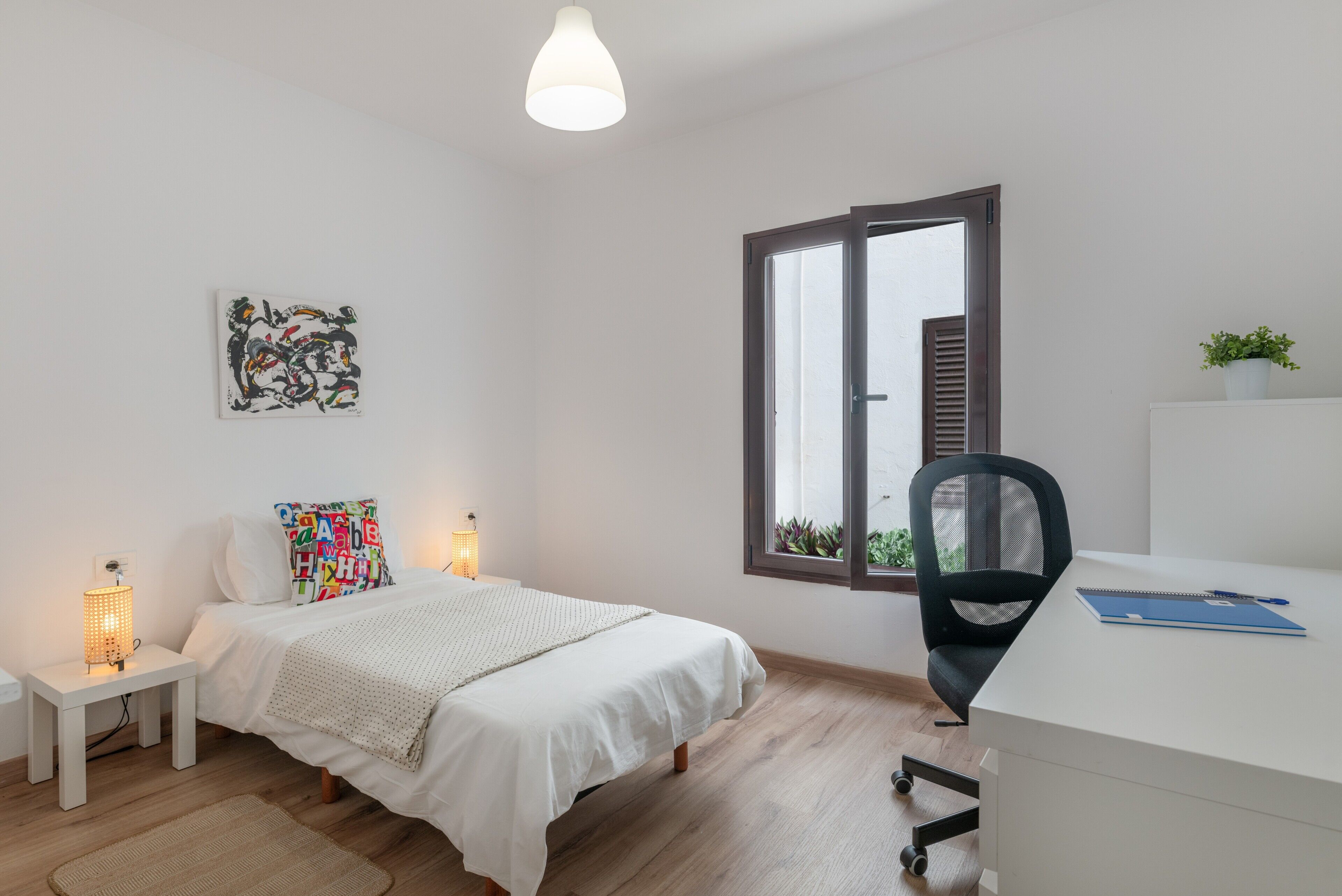 Een slaapkamer met een bed, nachtkastje, lamp, bureau, stoel, raam en kunst aan de muur.
