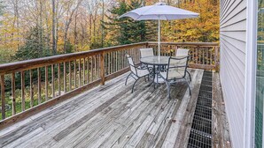 Een houten terras met een tafel, stoelen, een metalen hekwerk en uitzicht op herfstbomen.