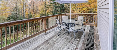 A wooden deck with a table and chairs, a metal railing, and a view of autumn trees.