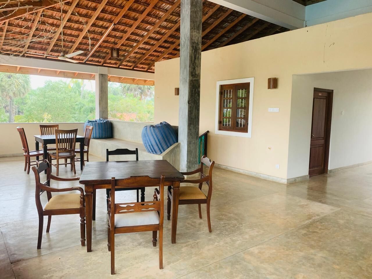 A spacious room with a thatched roof, a dining table with chairs, and a large window with a view of greenery outside.