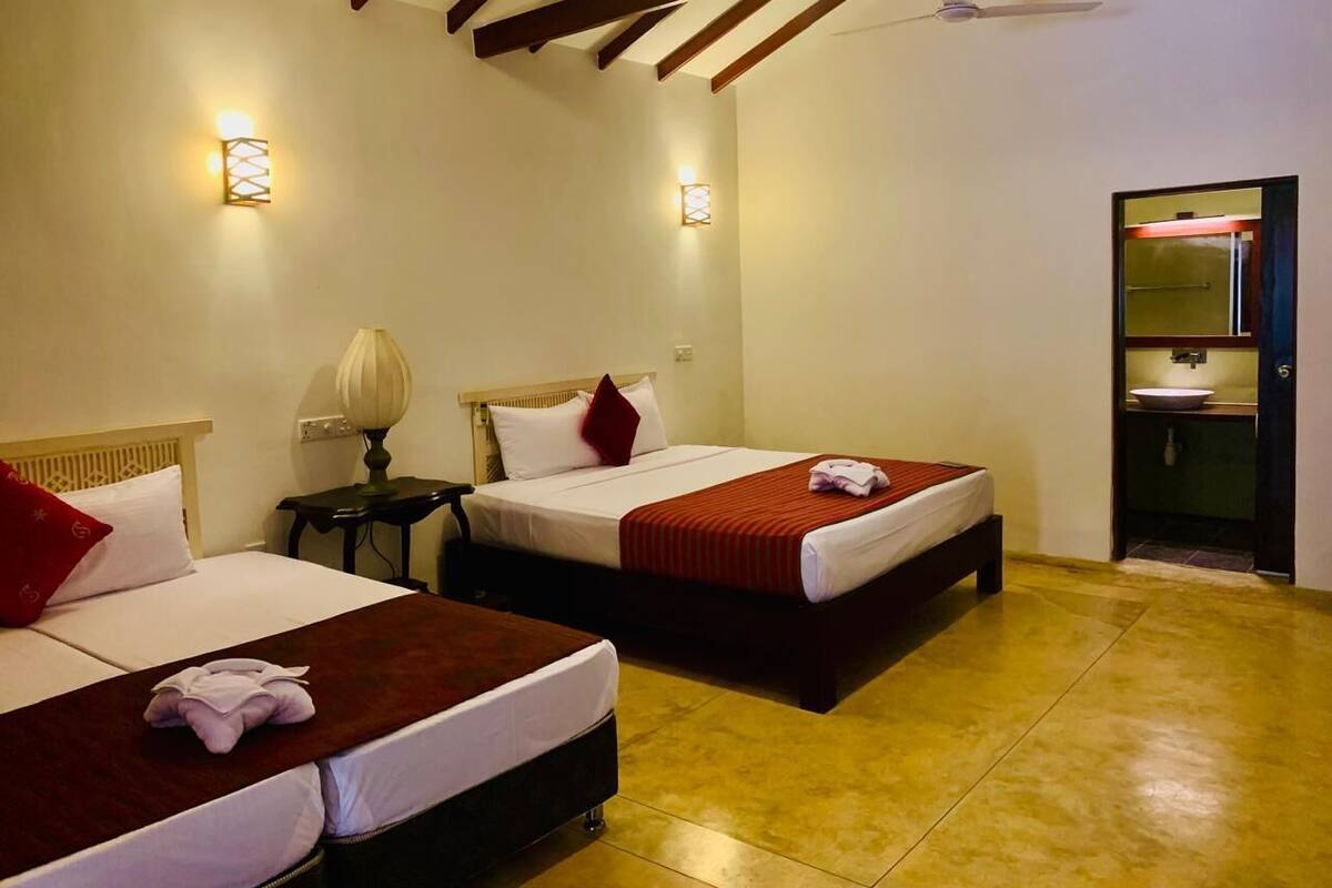 A hotel room with two beds, a nightstand with a lamp, a bathroom with a sink and mirror, and a ceiling fan.