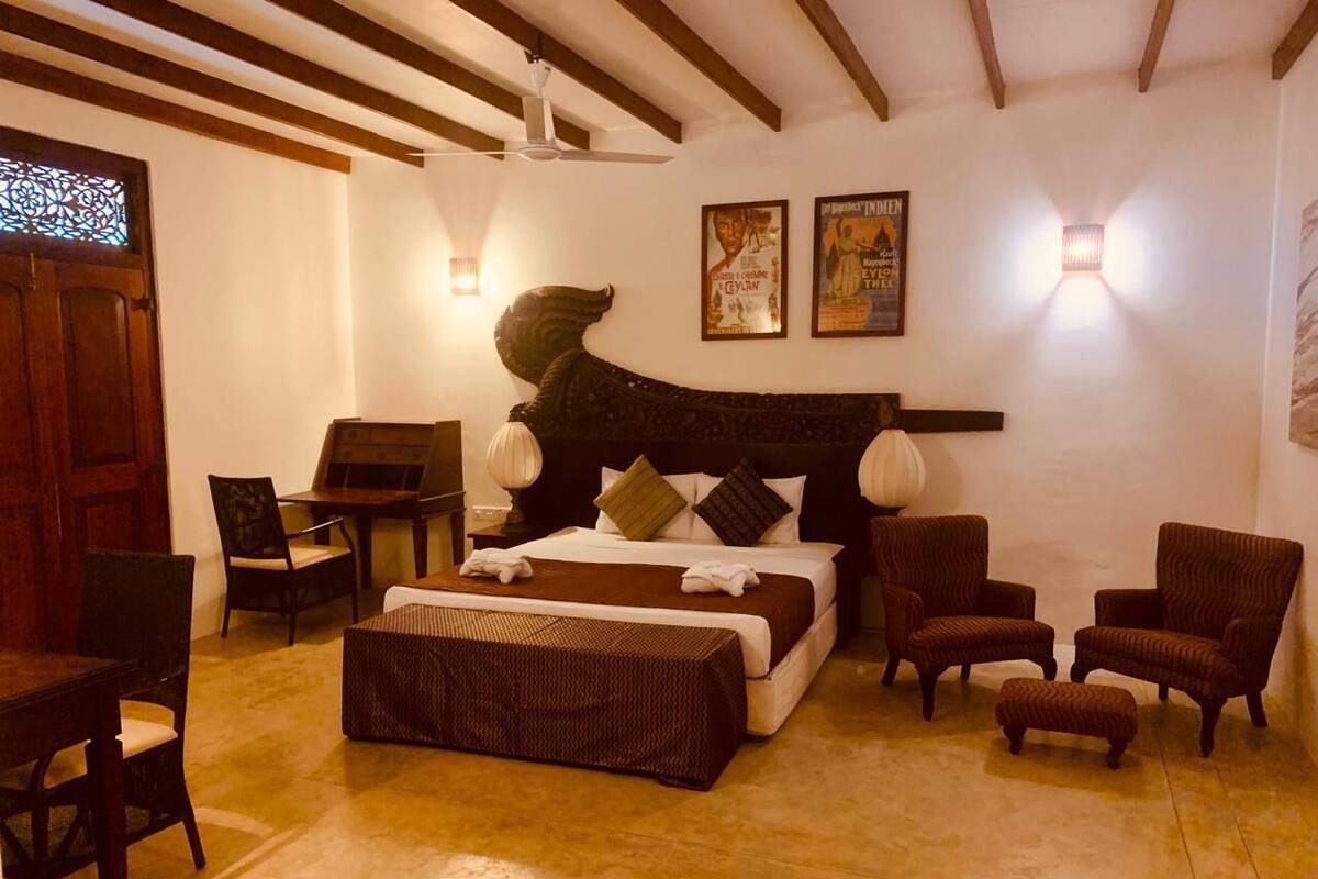 A room with a bed, two armchairs, a small table, and a chair. There are two wall-mounted lights and a poster on the wall.