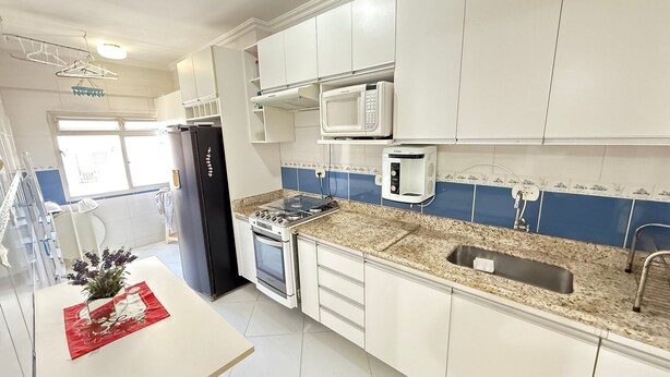 A modern kitchen with white cabinets, a black refrigerator, a gas stove, a microwave, and a sink. - Pé na Areia Praia do Canto do Forte IV (Praia Grande)