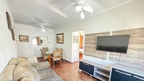A living room with a sofa, TV, and dining area. - Pé na Areia Praia do Canto do Forte IV (Praia Grande)