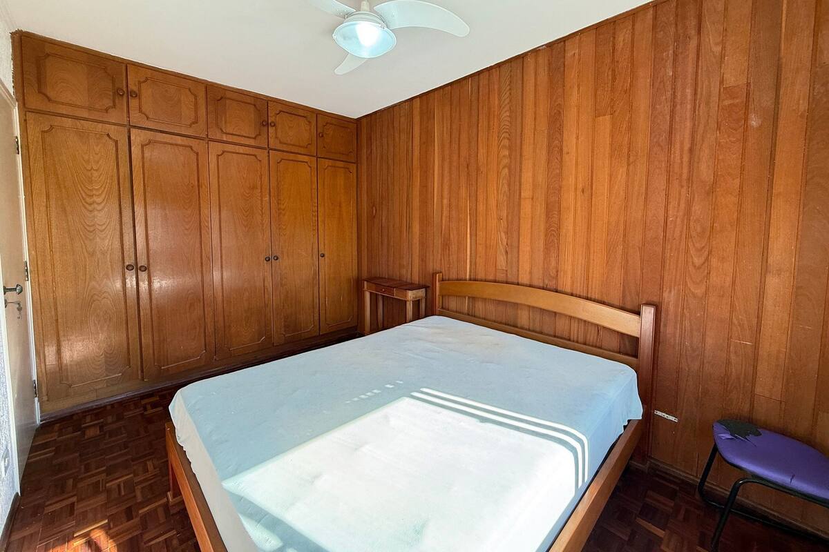 A bedroom with wooden paneling, a bed with a white mattress, a ceiling fan, and a small wooden nightstand.