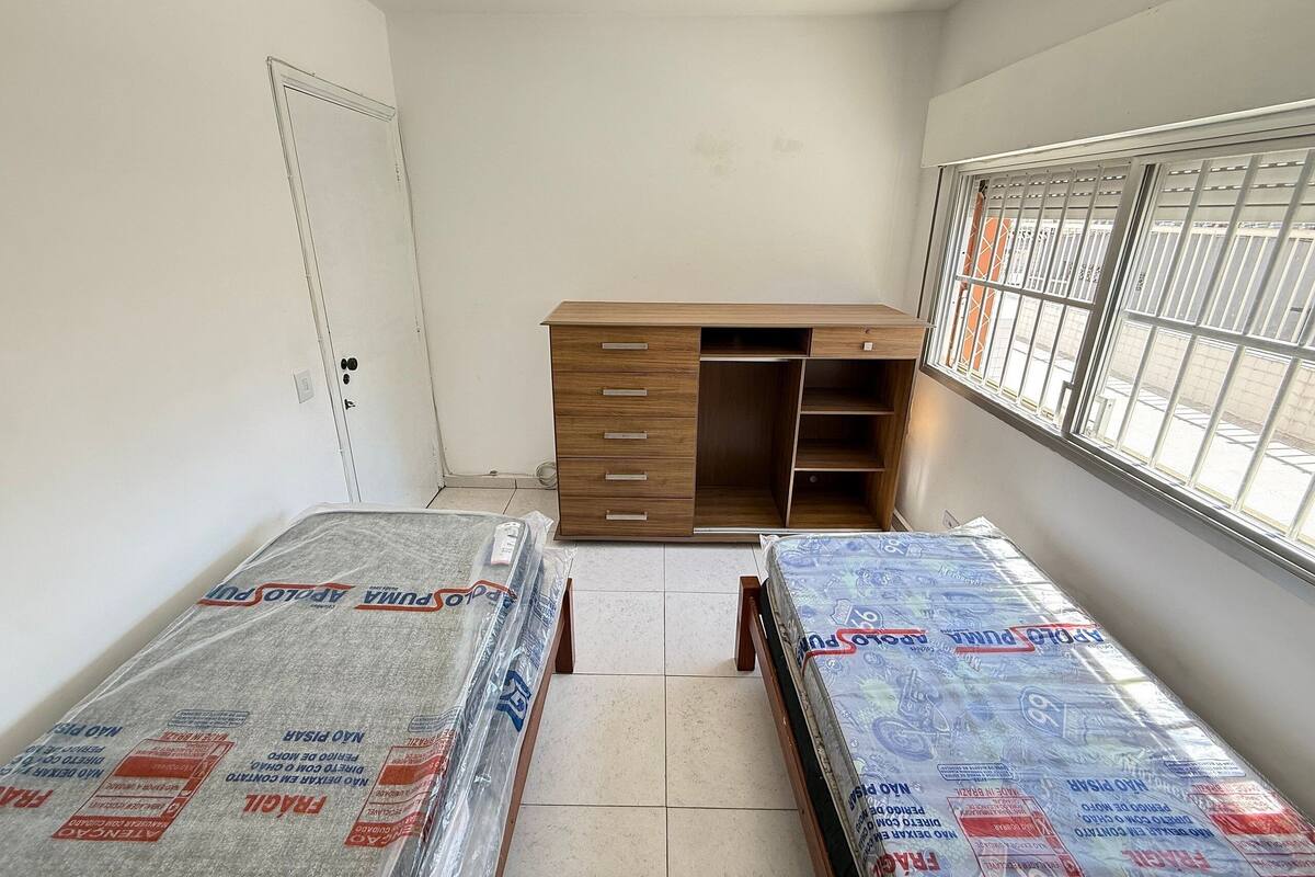 A room with a wooden wardrobe, two beds with mattresses, a window with bars, and a door.