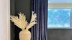 En trækommode med flettede skuffer, en vase med tørret pampasgræs, og et vindue med udsigt til grøn vegetation og lyserøde blomster.
