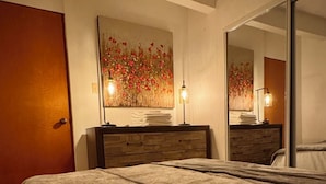 A bedroom with a large bed, a wooden dresser, a mirror, and a wall art piece.