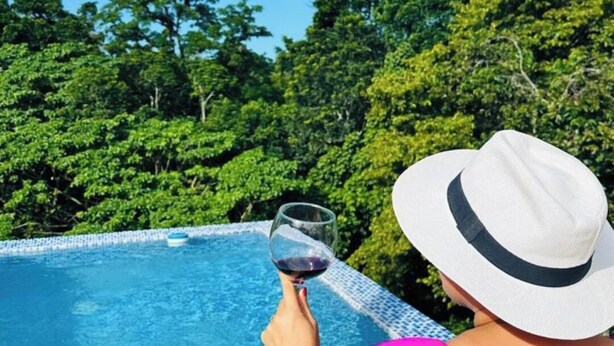Eine Person in einem rosa Badeanzug und einem weißen Hut sitzt an einem Pool mit klarem, blauem Wasser, umgeben von üppigem Grün und einem strahlend blauen Himmel.