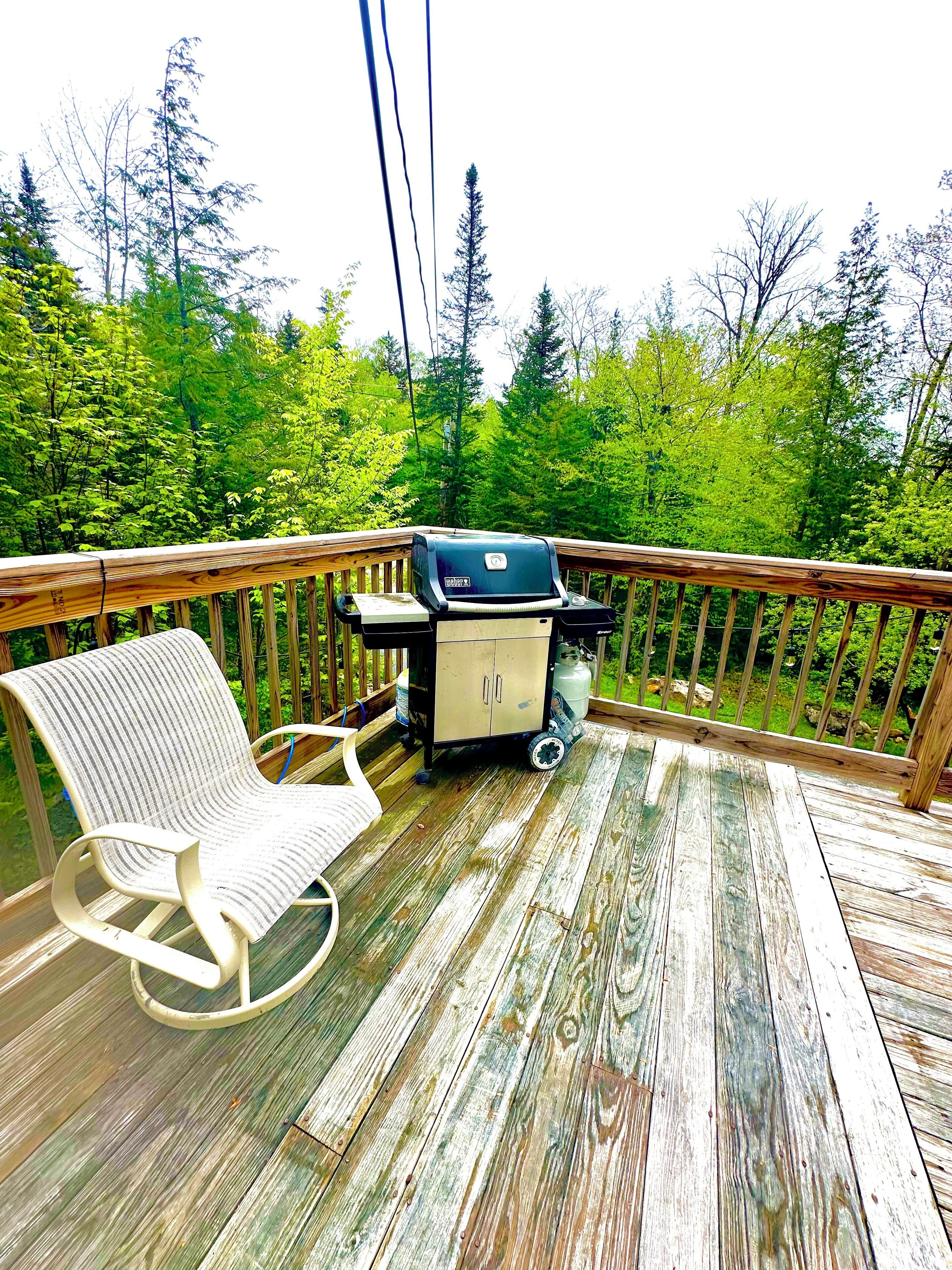 A wooden deck with a grill, a cushioned chair, and a view of a forest.