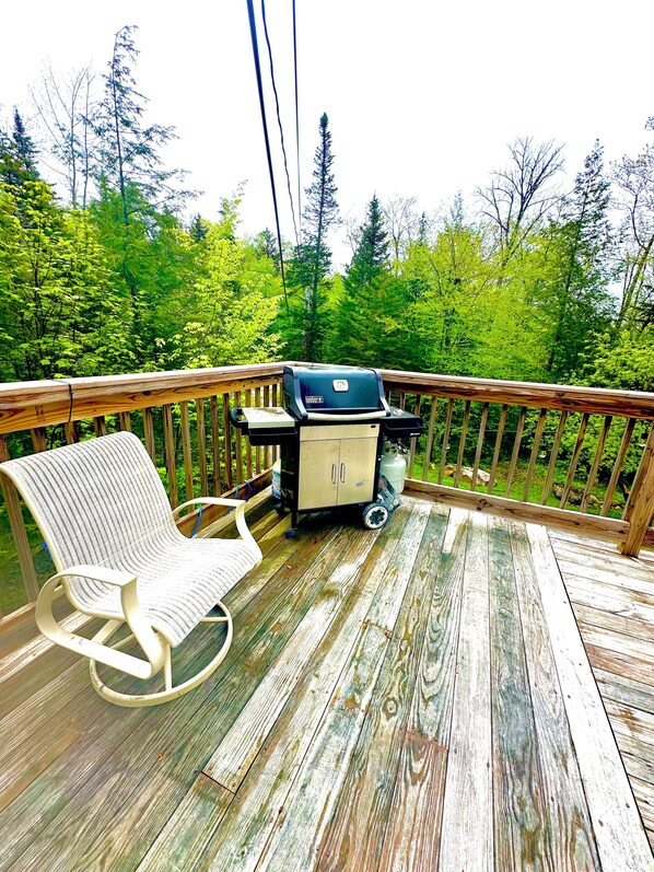 Una terraza de madera con una parrilla, una silla acolchada y vistas a un bosque.