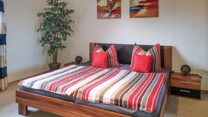 Una cama con una colcha a rayas, dos almohadas rojas y un cabecero de madera. Hay una planta en maceta a la izquierda de la cama y una mesita con una lámpara a la derecha.