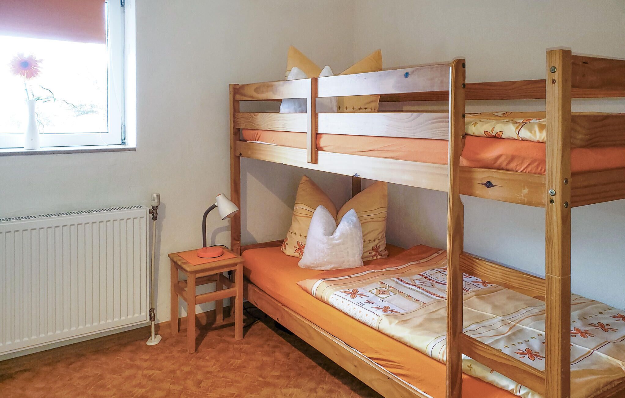 Ein Schlafraum mit Etagenbetten, einem Heizkörper, einem Schreibtisch mit Lampe und einem Fenster mit Blumen.