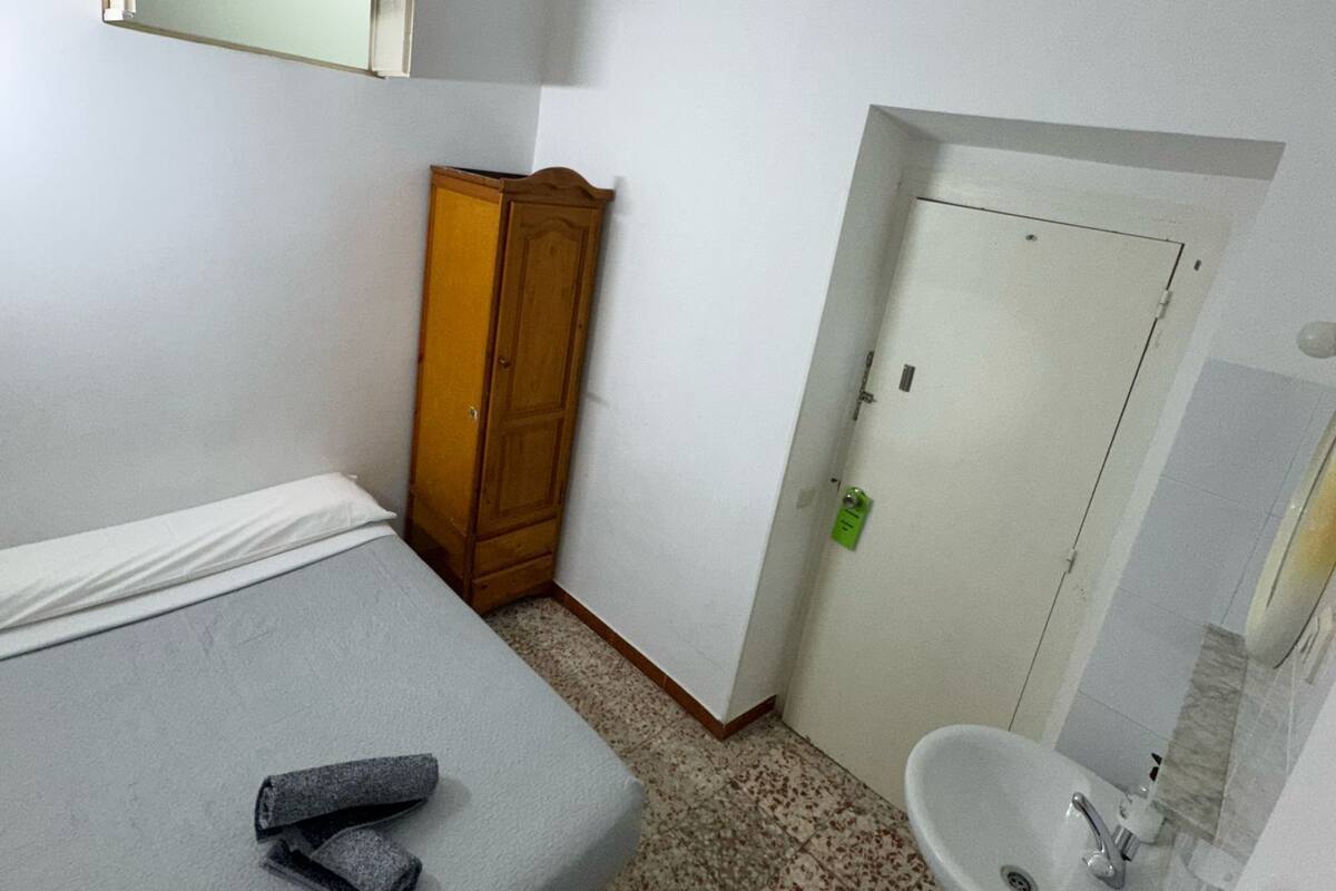 Een kleine, schone kamer met een bed, een houten garderobe, een badkamer met wastafel en spiegel, en een deur.