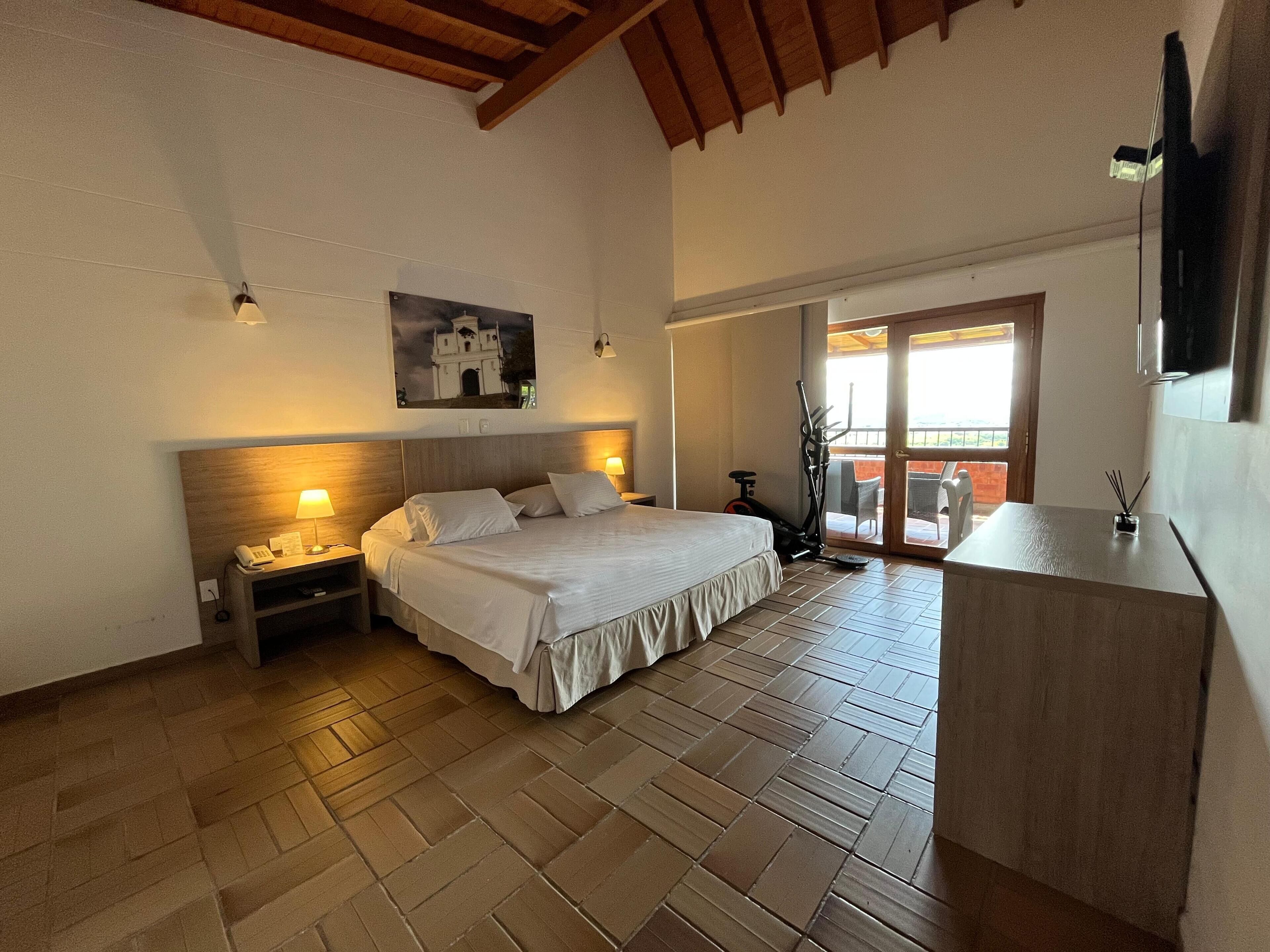 A bedroom with a bed, nightstands, a desk, and a view of the outside.