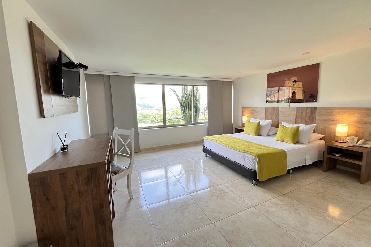 Une chambre d’hôtel comprenant un lit, des tables de chevet, un bureau, une chaise, un téléviseur fixé au mur et une fenêtre donnant sur un paysage verdoyant.