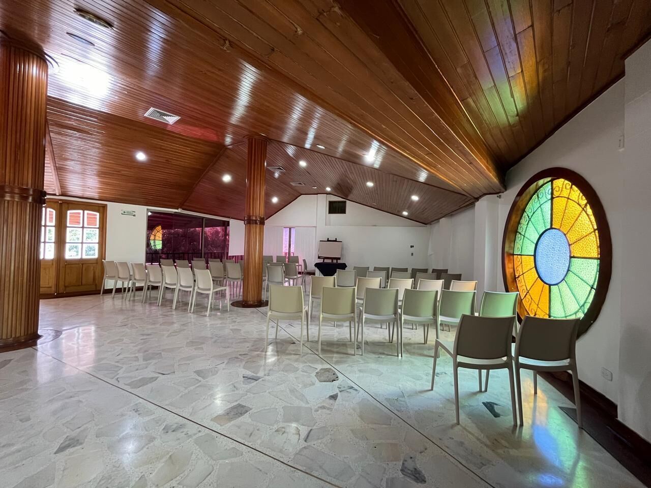 Un salón amplio con techo alto y un gran ventanal de vitrales. Filas de sillas están dispuestas para sentarse.