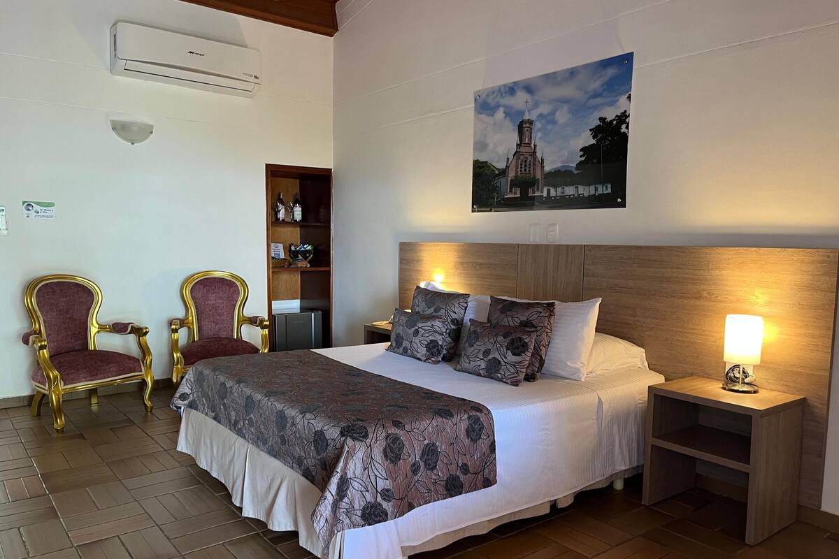 Une chambre d’hôtel avec un grand lit, deux fauteuils, une table de chevet en bois, un climatiseur fixé au mur, une étagère avec des bouteilles et un tableau représentant une église.