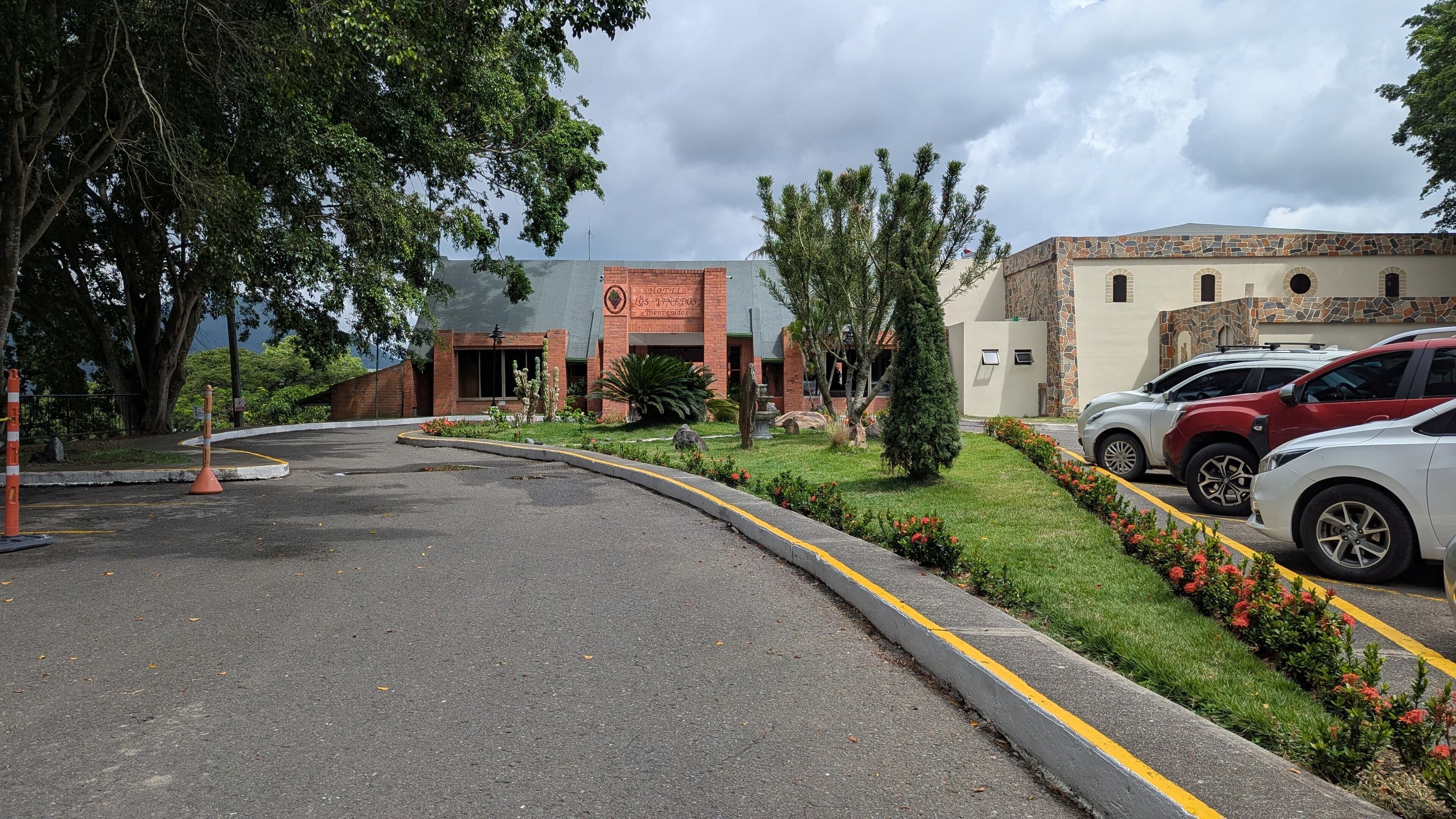 Entrada de un hotel con fachada de ladrillo, un área de estacionamiento con varios autos y un jardín ajardinado con arbustos y flores.