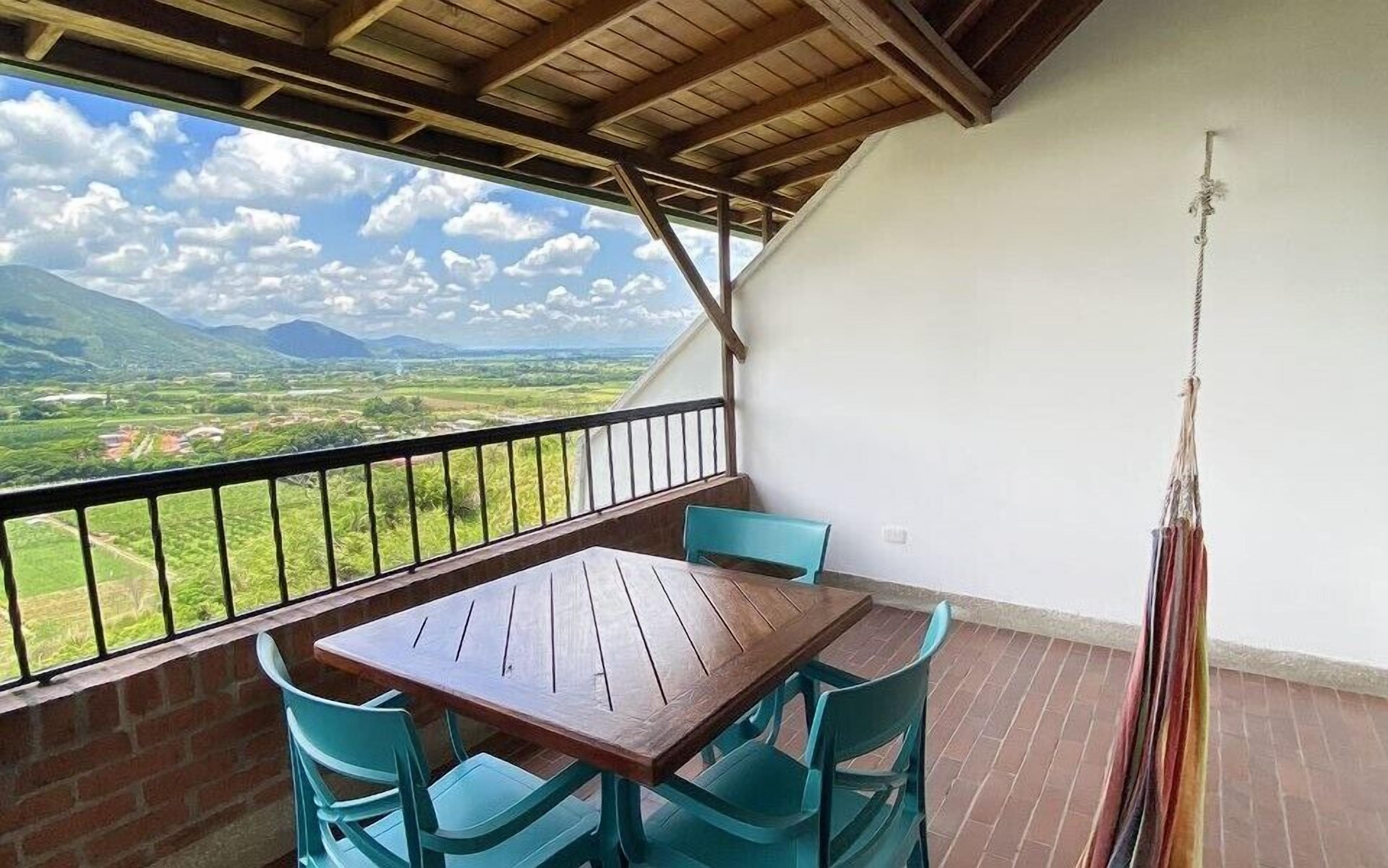 Un balcón con una mesa de madera y sillas turquesas, con vistas a un paisaje pintoresco con montañas y campos.