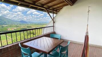 Un balcón con una mesa de madera y sillas turquesas, con vistas a un paisaje pintoresco con montañas y campos.