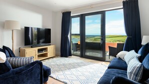 Un soggiorno moderno con una TV a schermo piatto su un mobile in legno, un divano con cuscini decorati e un'ampia porta scorrevole in vetro che dà su una terrazza esterna con vista panoramica.