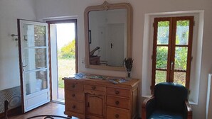 A room with a wooden dresser, a mirror, and a chair. There is a door leading outside, and a window with a view of greenery. - Mas des Louquiers (MONS)