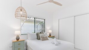 Une chambre à coucher comprenant un lit, deux tables de chevet, un luminaire suspendu, un ventilateur de plafond et une fenêtre avec des stores.