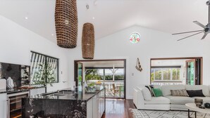 Une cuisine moderne avec un îlot central, un coin repas avec un canapé blanc et un tapis à motifs, et une vue sur la verdure par la porte ouverte.