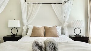 A neatly made bed with white linens, two beige pillows, and a pair of rolled towels. There are two bedside tables with lamps and a black frame. - 🤍Modern Alamo City Retreat🤍 (San Antonio)