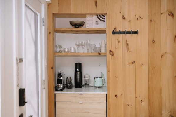 A wooden cabinet with shelves holding glassware, a coffee maker, and a kettle.