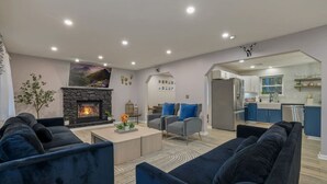 A modern living room with a fireplace, blue sofas, and a kitchen in the background.