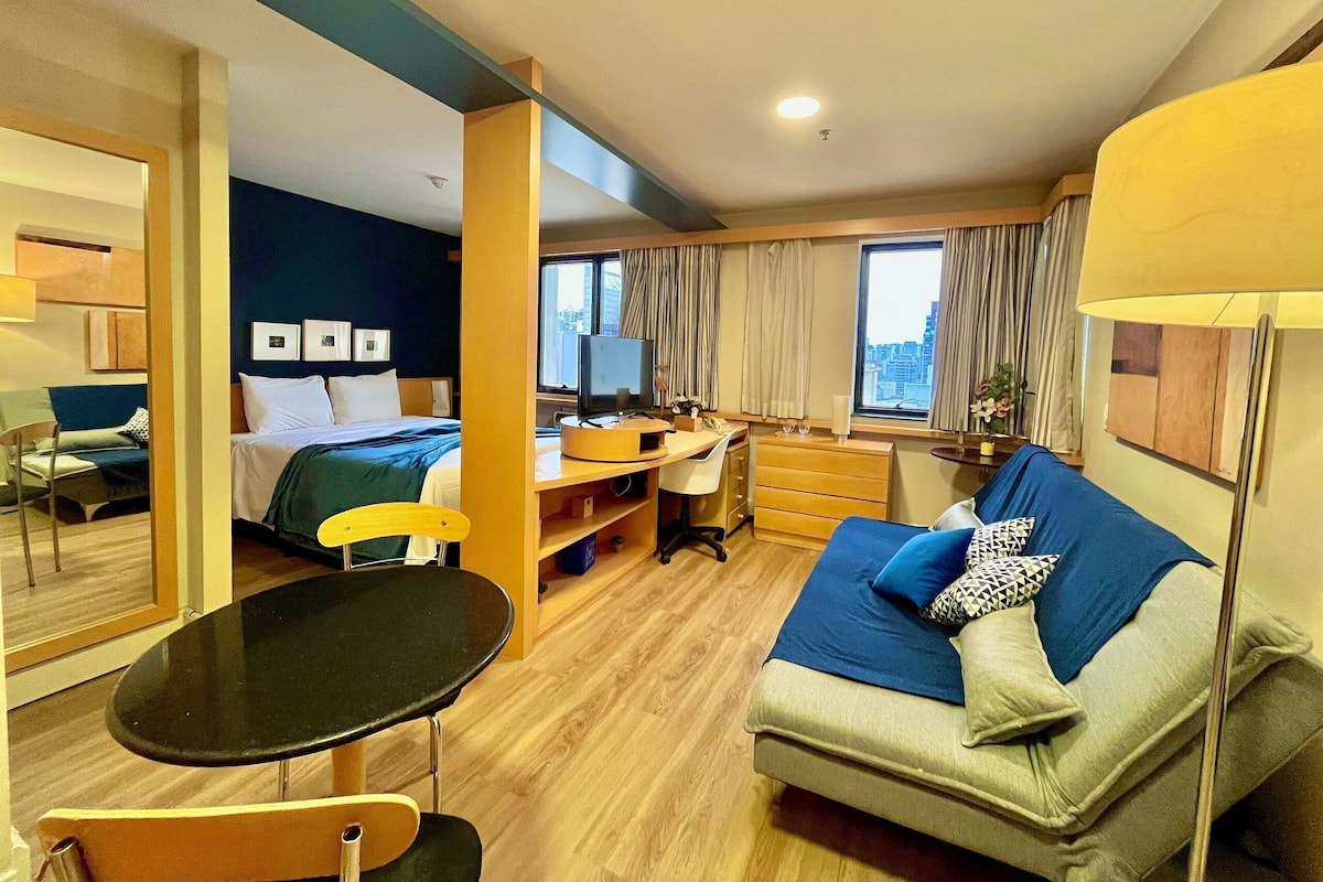 Habitación de hotel con cama, escritorio con computadora, sofá, mesita y ventana con cortinas.