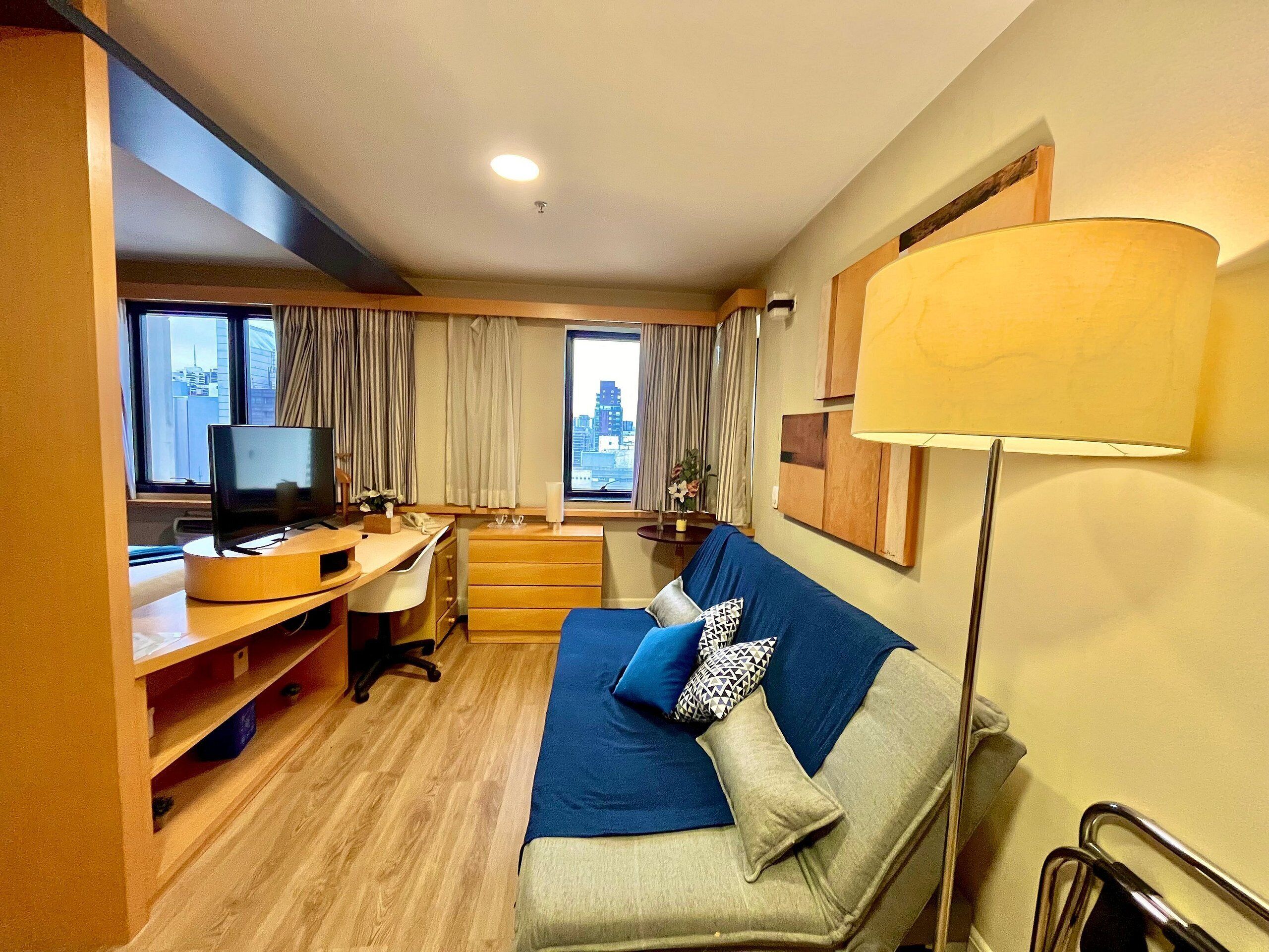 Ein Hotelzimmer mit Bett, Schreibtisch, Sessel, Fernseher und Fenstern mit Vorhängen.