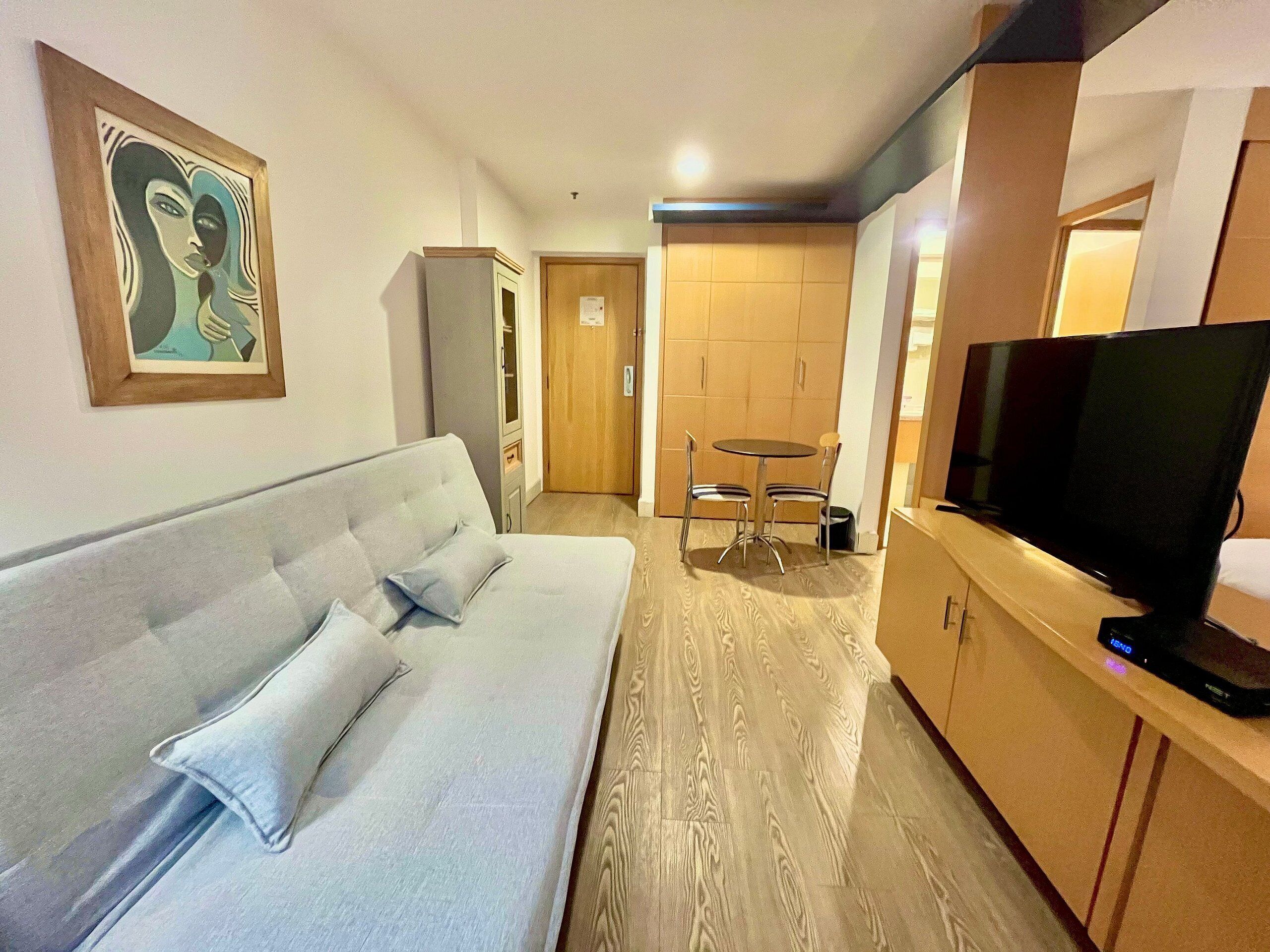 A modern hotel room with a grey sofa, a wooden cabinet, a television, and a dining area with a table and chairs.