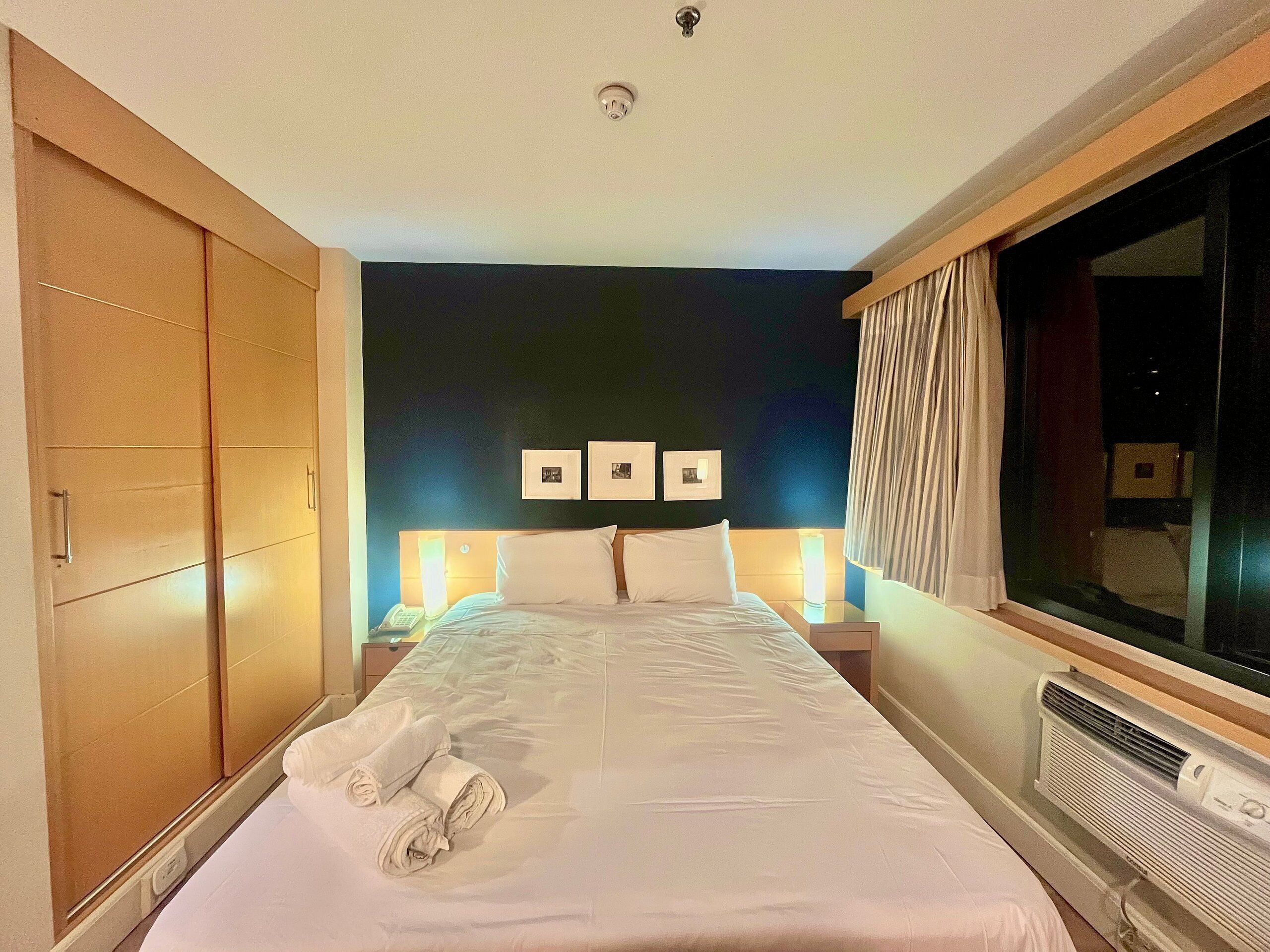 A hotel room with a large bed, a wardrobe, a bedside table with a lamp, and a window with curtains.