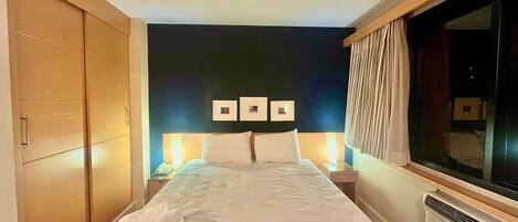 A hotel room with a large bed, a wardrobe, a bedside table with a lamp, and a window with curtains.