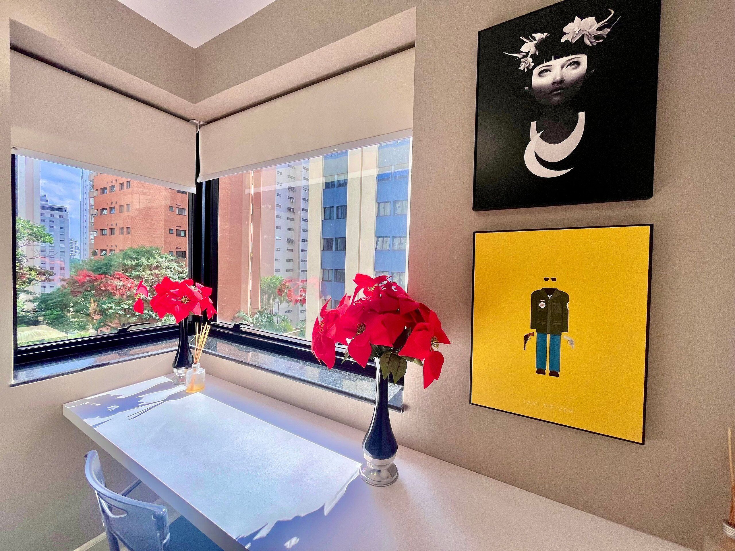 A hotel room with a large window, a table with a vase of red flowers, and two framed artworks on the wall.
