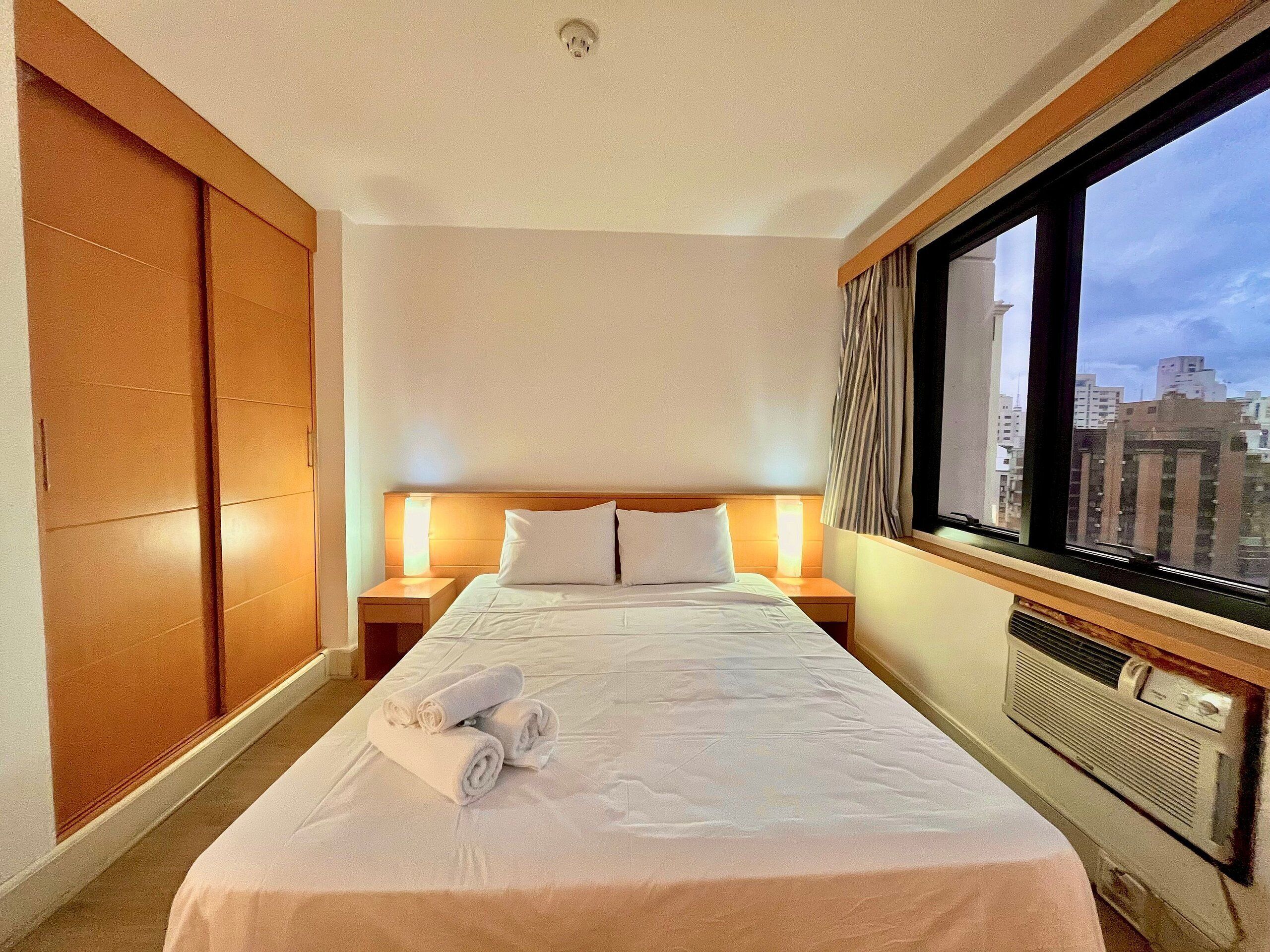 A hotel room with a bed, two bedside tables, a window with a city view, and a wardrobe.