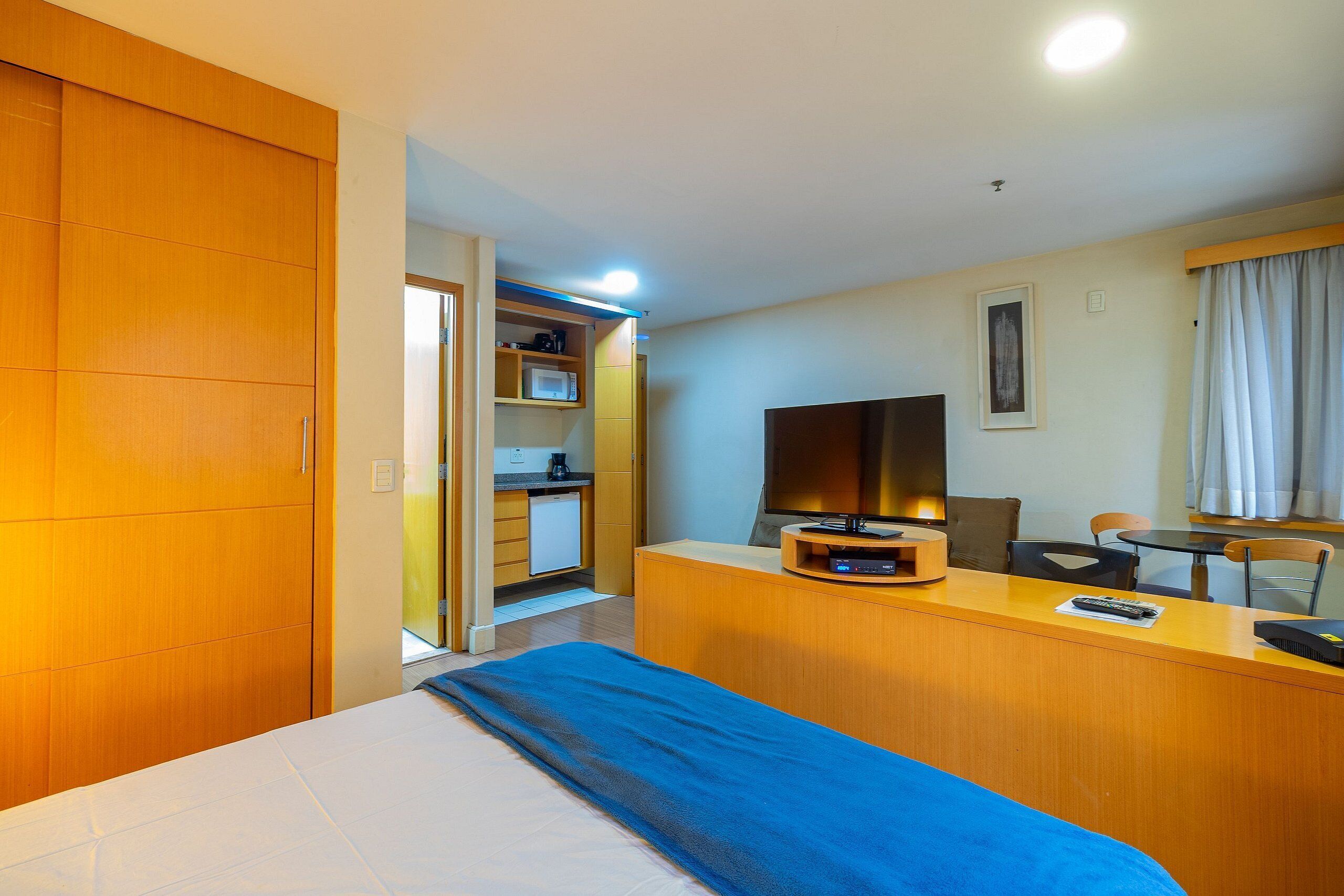 A hotel room with a bed, a desk with a TV, a chair, and a small kitchenette.