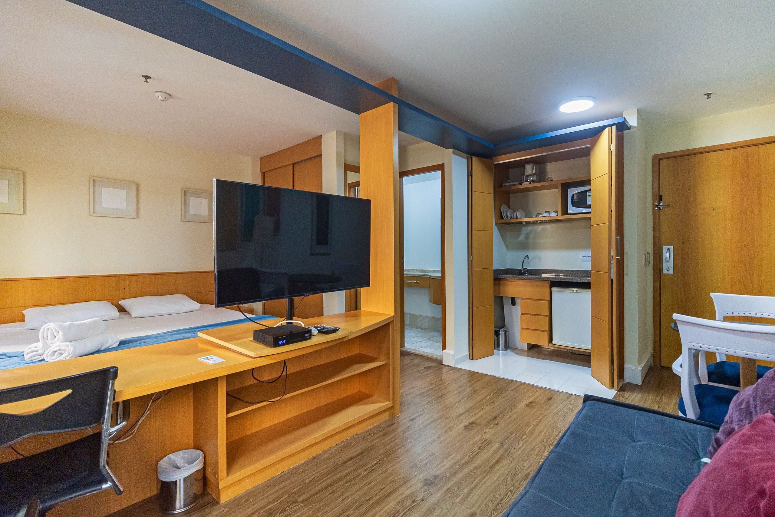 Habitación de hotel pequeña con cama, televisor, escritorio y kitchenette.