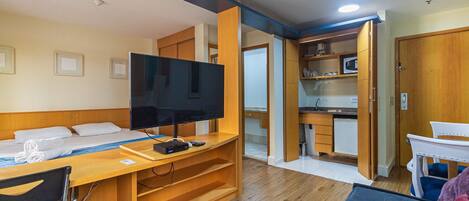 Habitación de hotel pequeña con cama, televisor, escritorio y kitchenette.