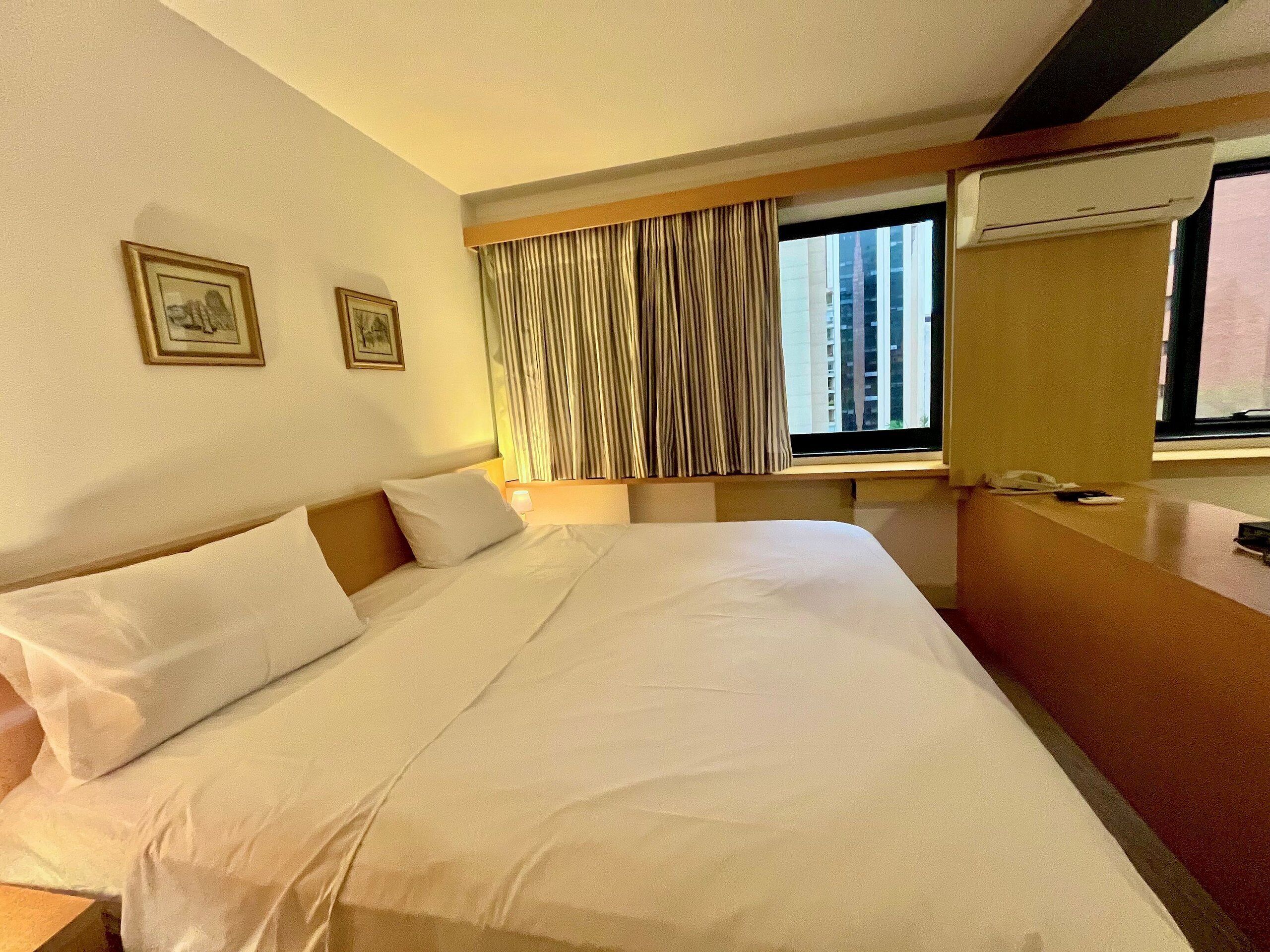 Una habitación de hotel con una cama, una mesita de noche, una ventana con cortinas y un aire acondicionado instalado en la pared.