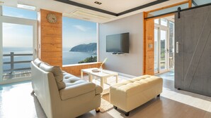 Un salon moderne avec un canapé, un pouf et une petite table. Un téléviseur est fixé au mur, et une grande fenêtre offre une vue sur la mer.