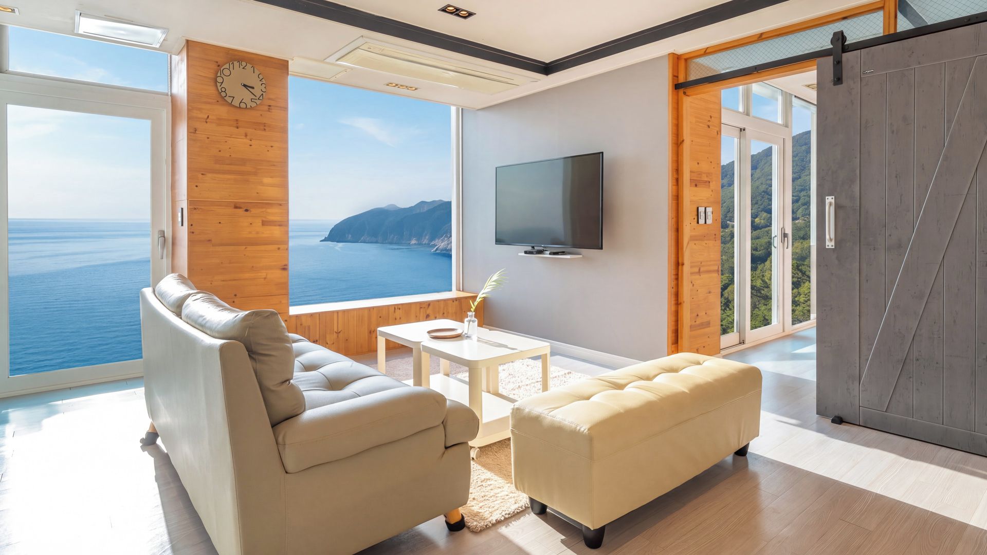 A modern living room with a large window overlooking the sea, a flat-screen TV mounted on the wall, and a sliding door leading to an outdoor area.