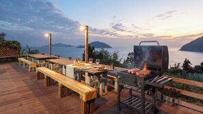 An outdoor dining area with wooden benches and tables, a barbecue grill, and a view of the sea. - Geoje Inbito Pension (Geoje)