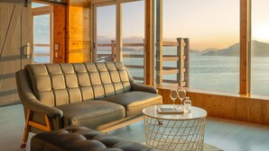 Une pièce moderne avec un canapé et un pouf en cuir, une table ronde avec des verres et un livre, et de grandes fenêtres offrant une vue sur la mer et les montagnes.