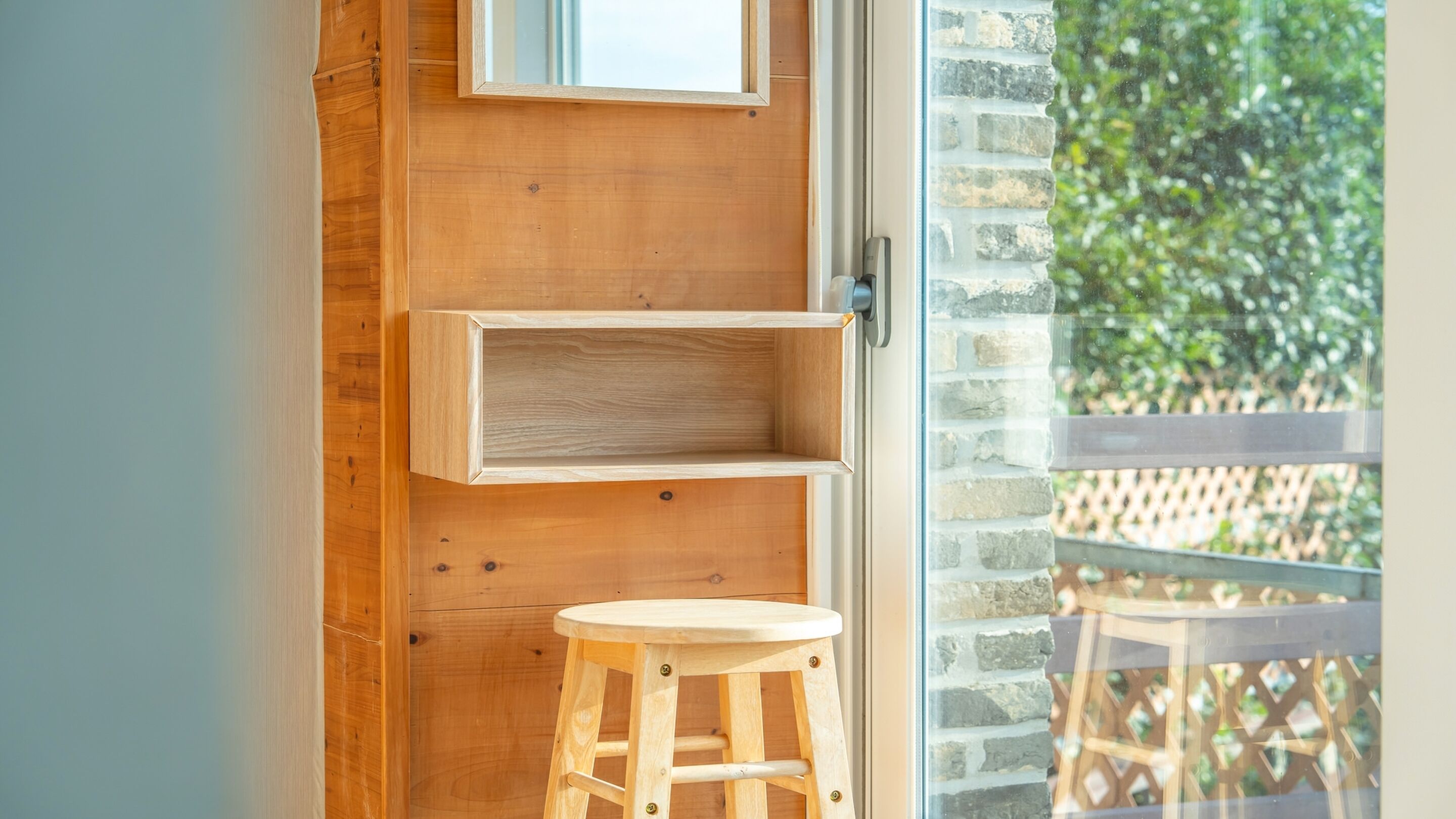 A wooden interior with a wooden stool, a shelf, and a door leading to a balcony with a view of greenery.