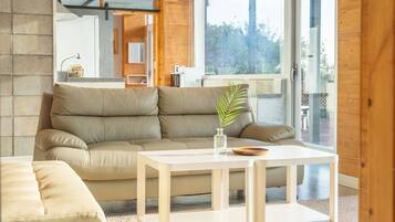 A modern living room with a beige leather sofa, a wooden coffee table, and a view of a kitchen and outdoor area through large windows.