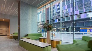 Eine moderne Lobby mit grünen Sitzgelegenheiten, einer großen Topfpflanze und Blick auf die Stadt durch große Fenster.